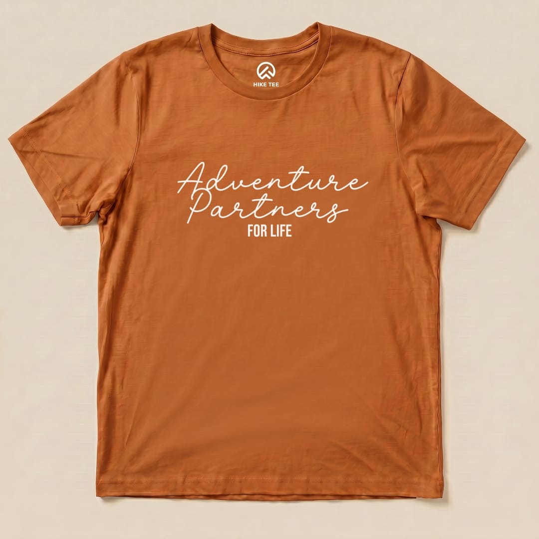 A flat graphic t-shirt in rust color featuring the phrase "Adventure Partners for Life" in a stylish font.
