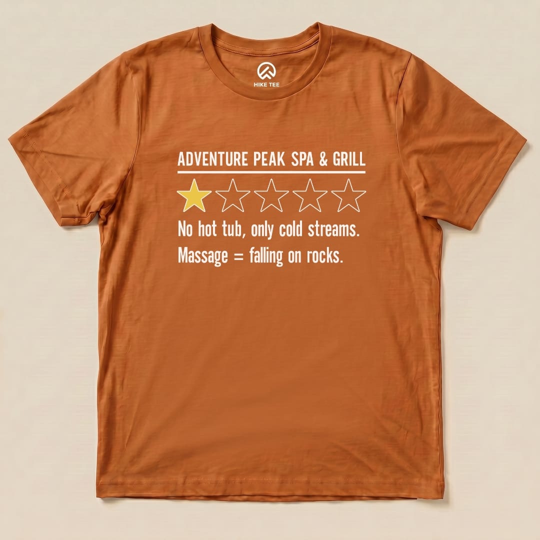 The graphic t-shirt features the humorous text "ADVENTURE PEAK SPA & GRILL," with one yellow star and a review about cold streams and rocky massages, labeled as the "Adventure Spa & Grill Review