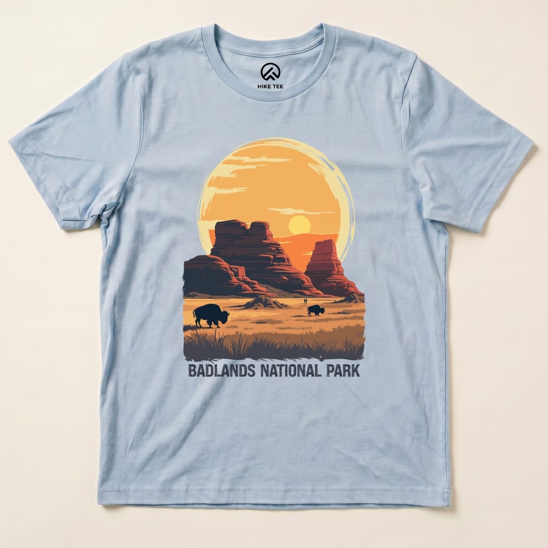 A light blue graphic t-shirt featuring a sunset over rock formations and bison, labeled "Badlands National Park Shirt.