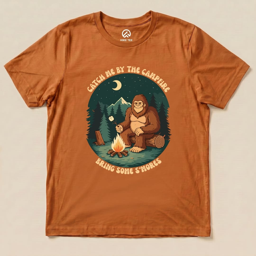 Orange graphic t-shirt featuring a Bigfoot character by a campfire, with the slogan "Catch Me By The Campfire Bring Some S'mores".