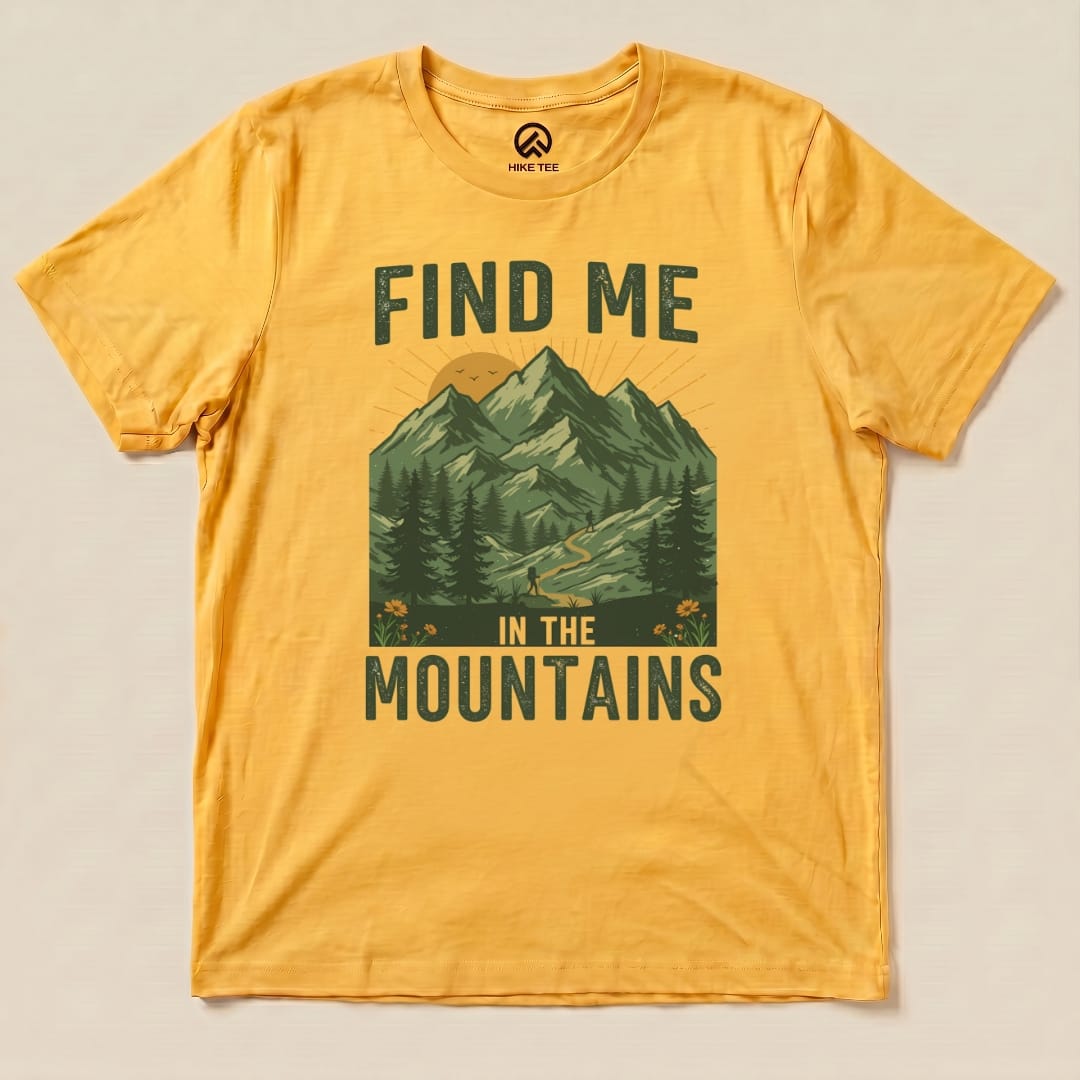 A graphic t-shirt in golden yellow featuring the phrase "Find Me In The Mountains" and a mountain landscape design.