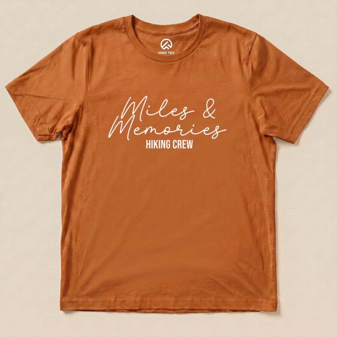 A flat view of an orange graphic t-shirt featuring the words "Miles & Memories" with "Hiking Crew" underneath.