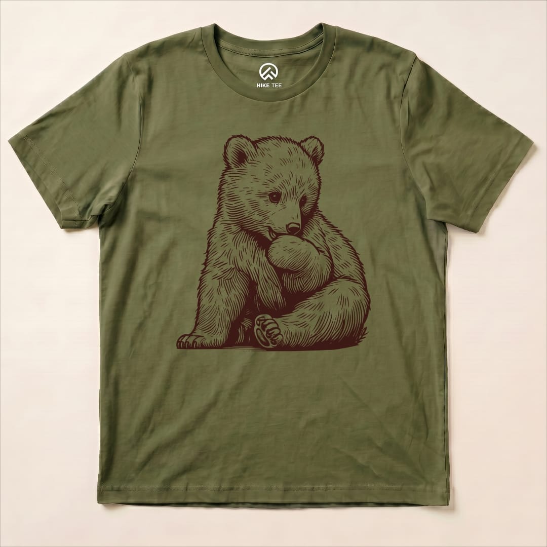 A graphic t-shirt featuring an adorable illustration of a bear cuddling a ball, in a muted olive green color, labeled as "Youth Bear Shirt.