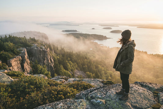Sunrise view from Cadillac Mountain in Acadia National Park with coastal fog, granite peaks, Atlantic Ocean, and lone hiker overlooking the landscape.