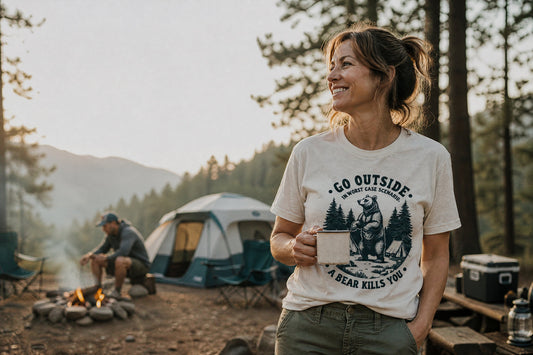 Woman wearing a bear wildlife graphic T-shirt enjoys a quiet morning at a forest campsite with coffee, tent, and camping gear in the background.