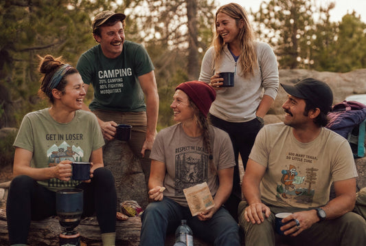 A mixed group of hikers at a campsite or trail stop, with some wearing funny hiking quote shirts and others in plain outdoor clothing.