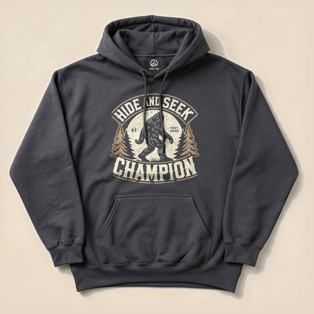 A charcoal hoodie featuring a graphic design of Bigfoot and the text "Hide and Seek Champion" surrounded by trees.