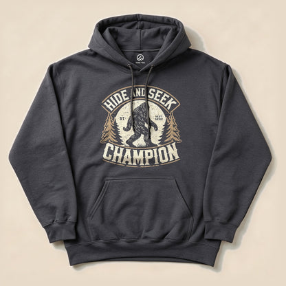 A charcoal hoodie featuring a graphic design of Bigfoot and the text "Hide and Seek Champion" surrounded by trees.