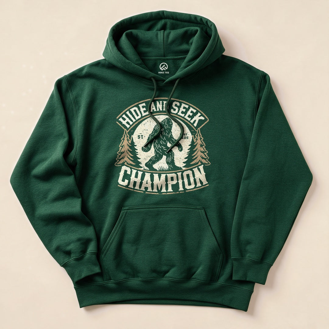 A dark green hoodie featuring a graphic of Bigfoot amid trees, with the text "Hide and Seek Champion" prominently displayed.