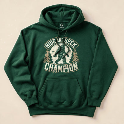 A dark green hoodie featuring a graphic of Bigfoot amid trees, with the text "Hide and Seek Champion" prominently displayed.