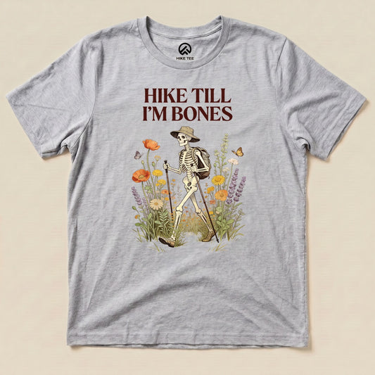 Athletic Heather Hike Till I'm Bones Shirt with a skeleton hiker walking through wildflowers graphic