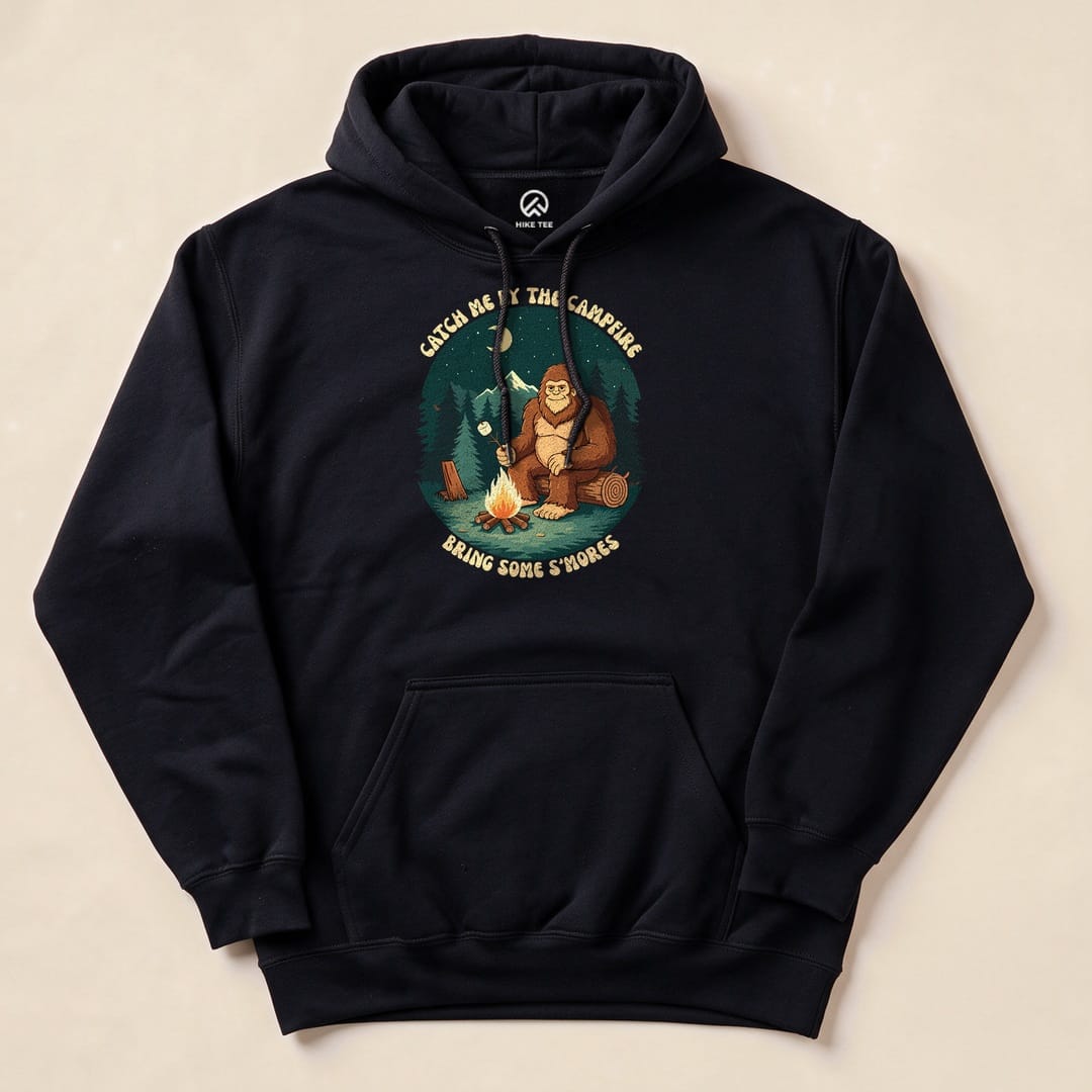A navy hoodie featuring a graphic of a bear by a campfire with the text "Catch Me By The Campfire Bring Some S'mores.