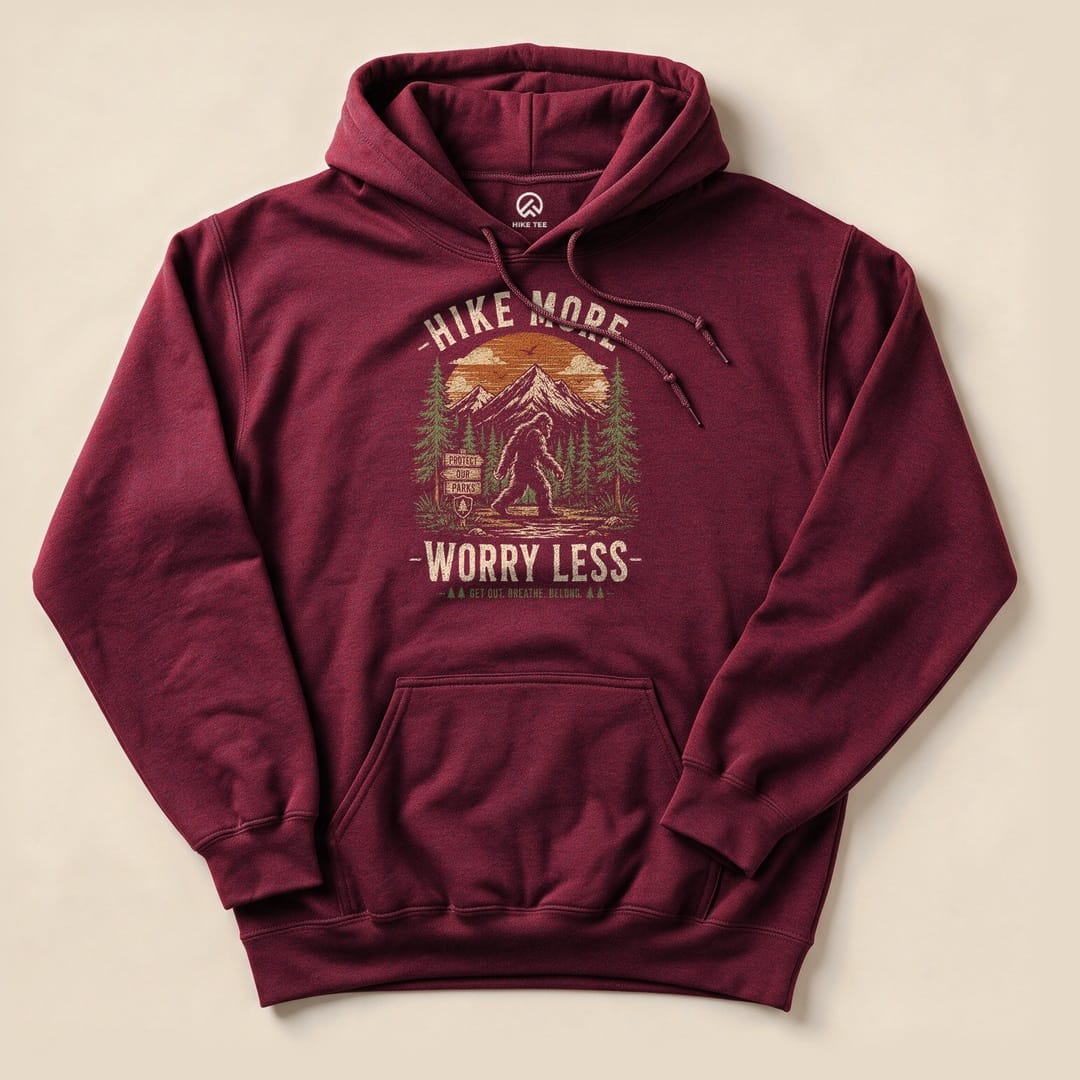A burgundy hoodie featuring a colorful graphic of Bigfoot amidst trees and mountains, with the text "Hike More, Worry Less.