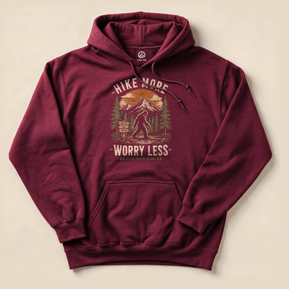 A burgundy hoodie featuring a colorful graphic of Bigfoot amidst trees and mountains, with the text "Hike More, Worry Less.