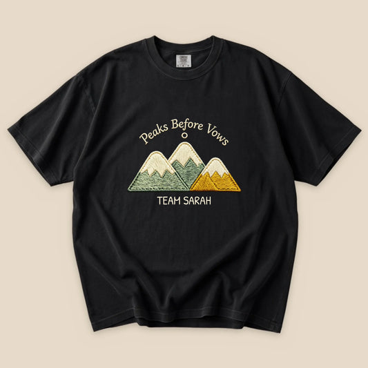 A black graphic t-shirt featuring an embroidered design of mountains with the text "Peaks Before Vows" and "TEAM SARAH" underneath.