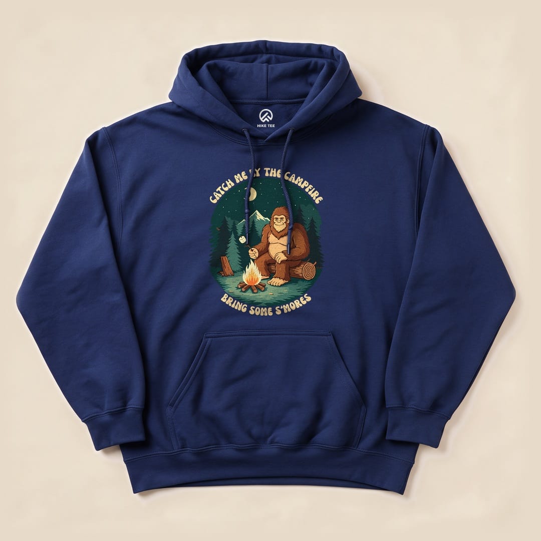 A navy graphic hoodie features a playful illustration of Bigfoot sitting by a campfire, with the text "Bring Some S'mores".
