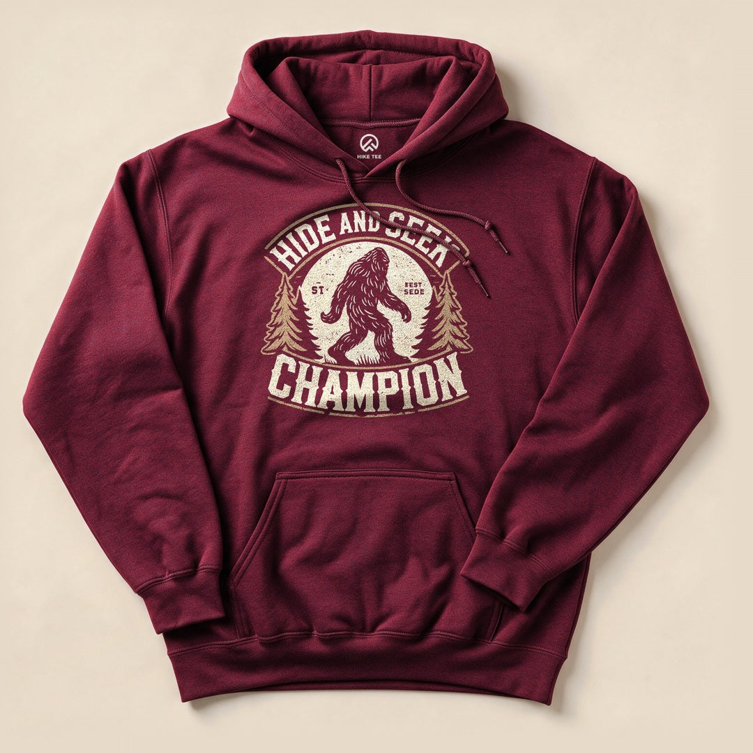 A maroon hooded sweatshirt featuring a graphic of Bigfoot with the phrase "Hide and Seek Champion" above and trees on the sides.
