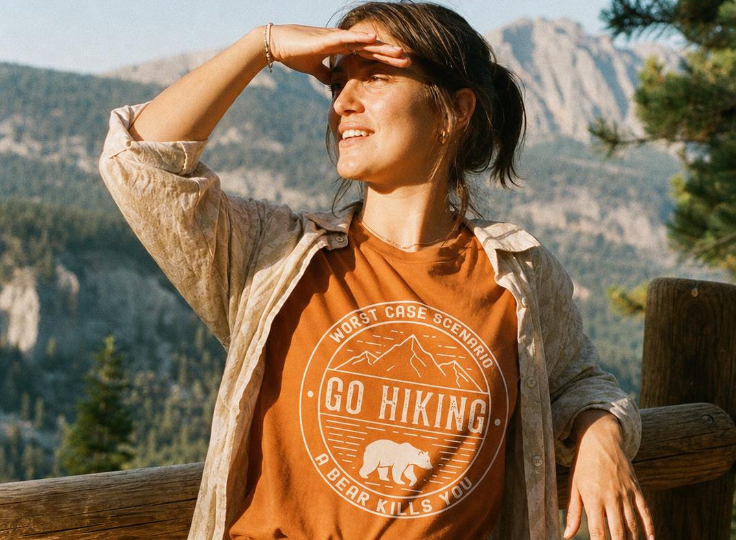 A woman outdoors in front of mountains and trees, wearing an orange graphic T-shirt under a light beige button-up shirt and denim shorts. The T-shirt has a circular hiking-style print with mountains, a bear, and the text 'GO HIKING' and 'MOST CASE SCENE AHEAD' plus 'BEAR KILLS YOU'.