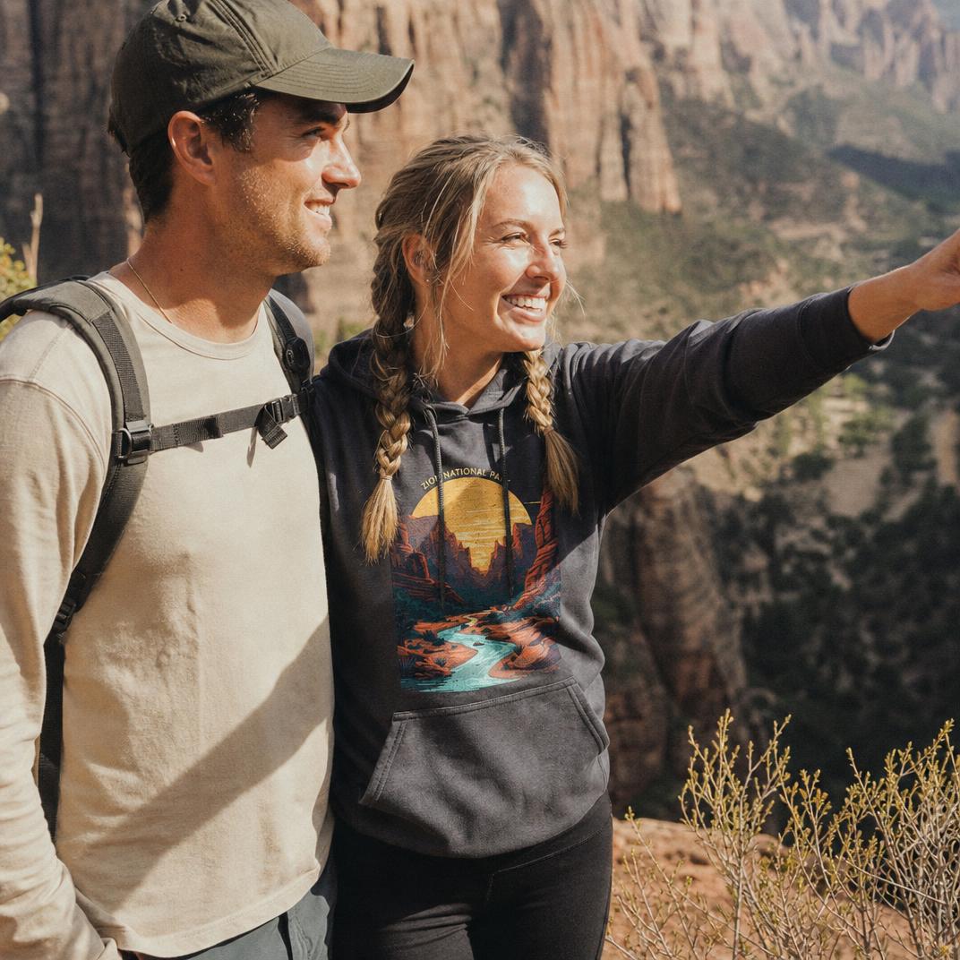 A smiling woman wearing a dark gray graphic hoodie with a sunset landscape print and black pants stands on a mountain trail next to a man in a beige long-sleeve shirt and green cap, pointing toward a scenic canyon view.
