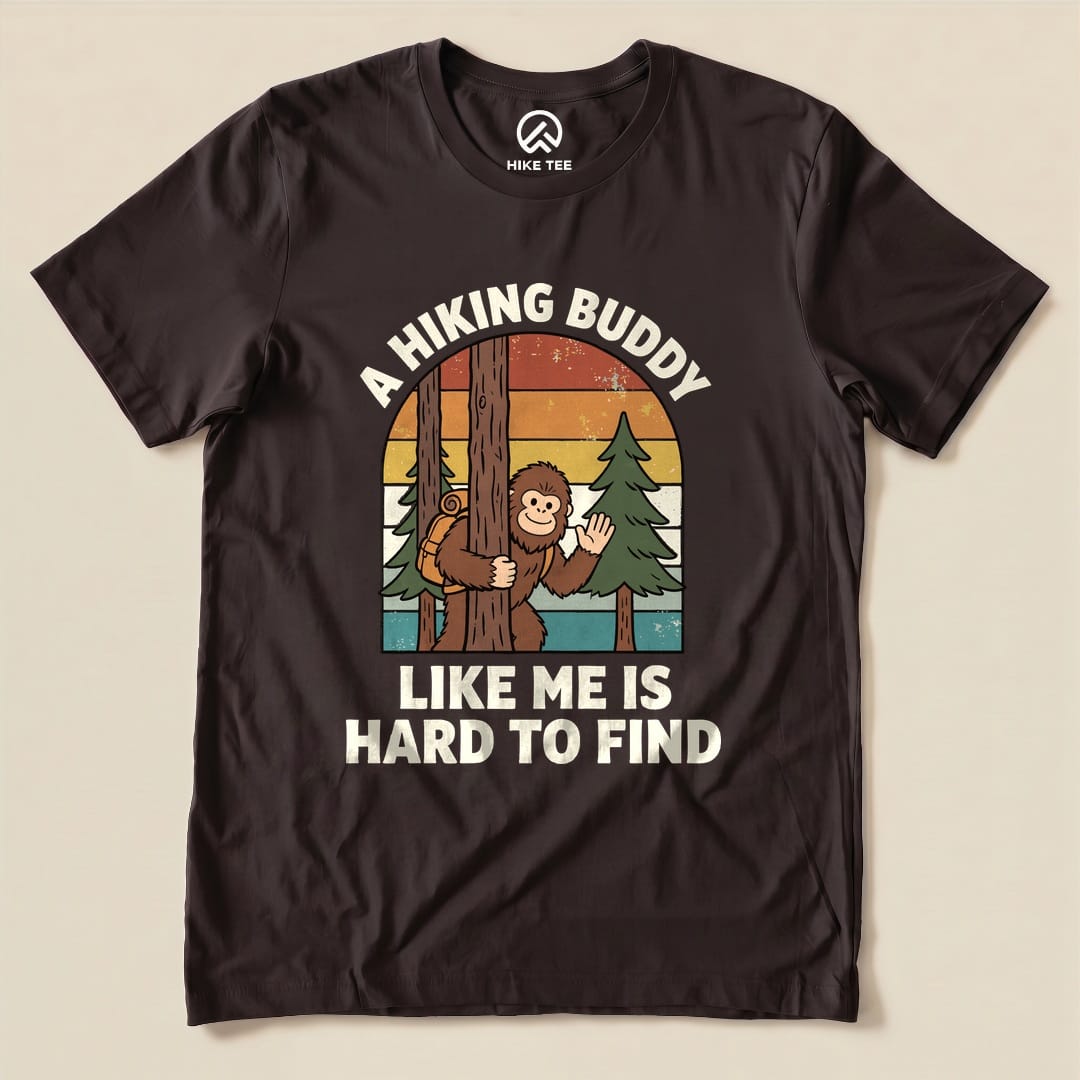 A graphic t-shirt featuring a playful design of a cartoon monkey peeking from behind a tree with the text "A Hiking Buddy Like Me Is Hard To Find.
