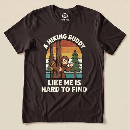 A graphic t-shirt featuring a playful design of a cartoon monkey peeking from behind a tree with the text "A Hiking Buddy Like Me Is Hard To Find.