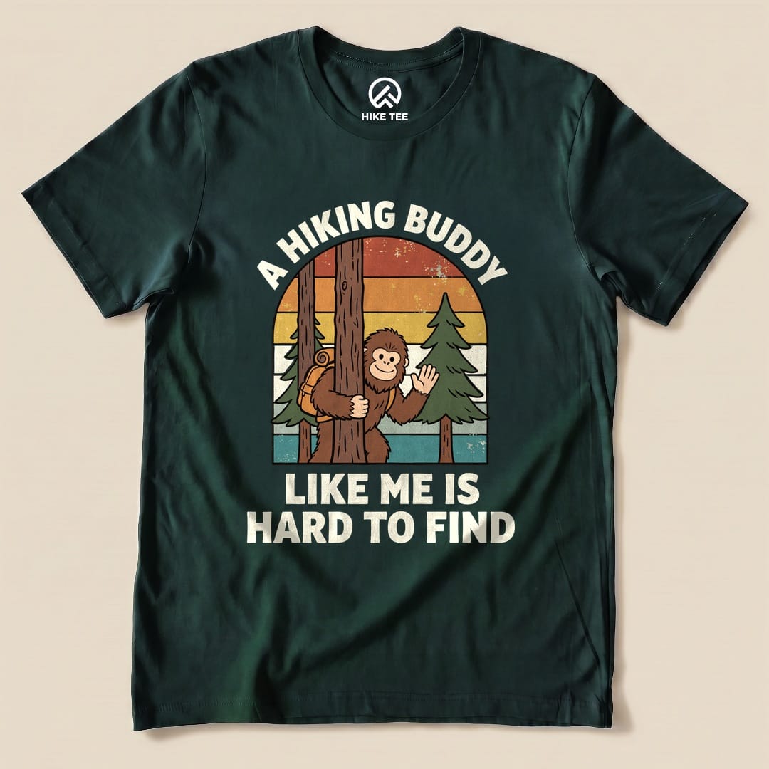 The graphic t-shirt features a playful design with a cartoon monkey peeking from behind a tree and the slogan "A Hiking Buddy Like Me Is Hard To Find.