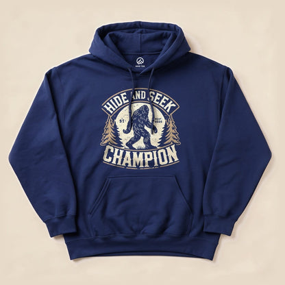 A navy blue hoodie featuring a graphic design of Bigfoot and the text "Hide and Seek Champion" surrounded by pine trees.