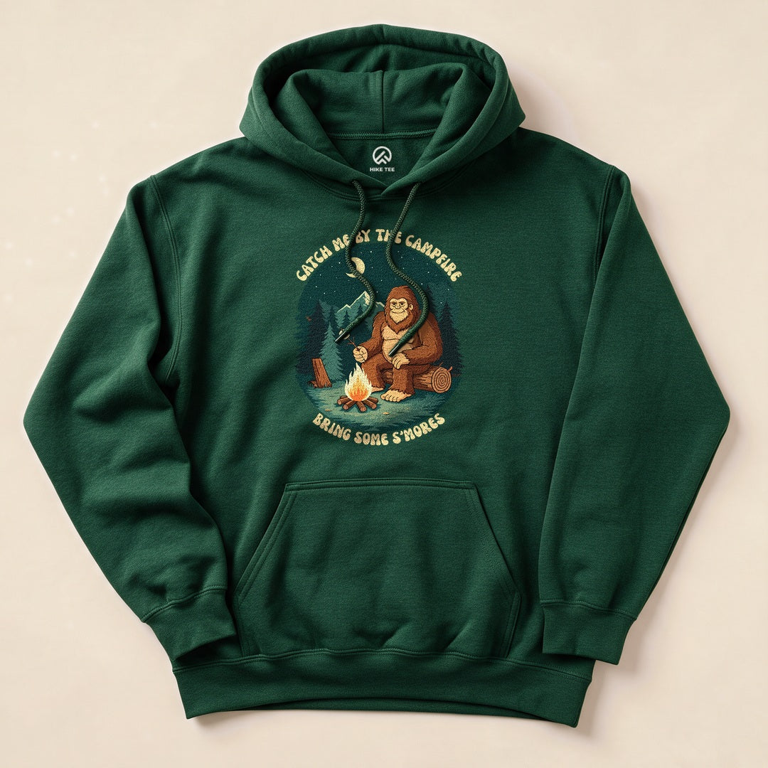 A green hoodie featuring a graphic of a Sasquatch by a campfire with the text "Catch Me By The Campfire, Bring Some S'mores".