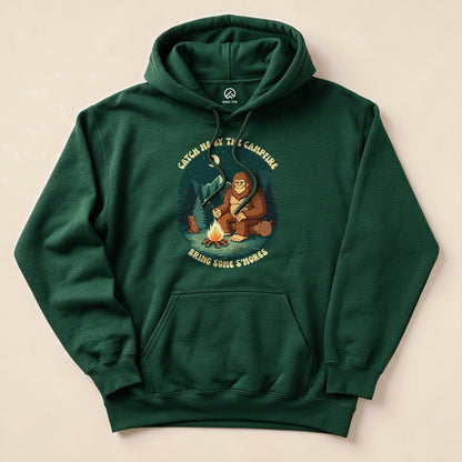 A green hoodie featuring a graphic of a Sasquatch by a campfire with the text "Catch Me By The Campfire, Bring Some S'mores".