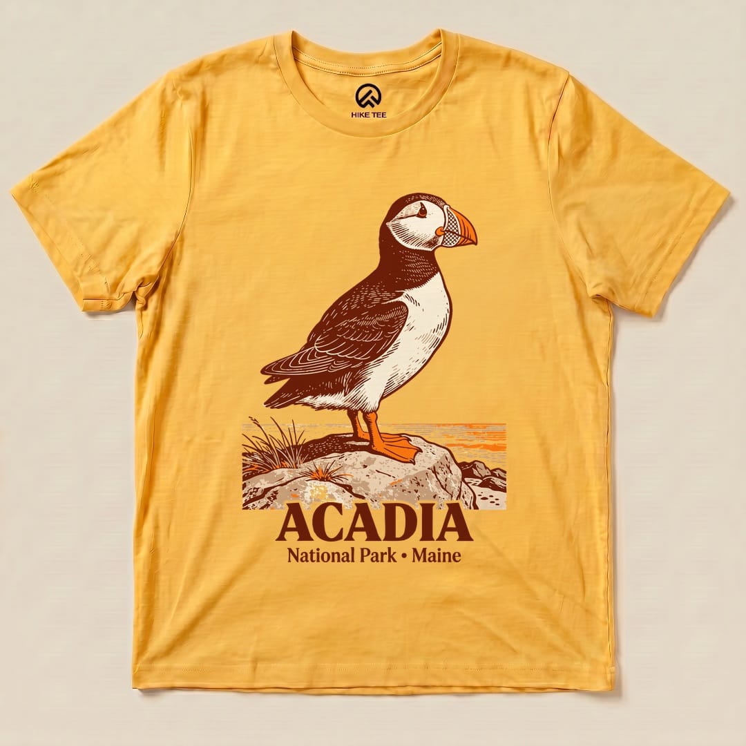 A graphic t-shirt featuring a puffin standing on a rock with the text "Acadia National Park • Maine" below.