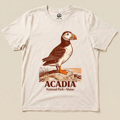 A graphic t-shirt featuring a puffin standing on rocks, with "ACADIA National Park • Maine" printed below the design.