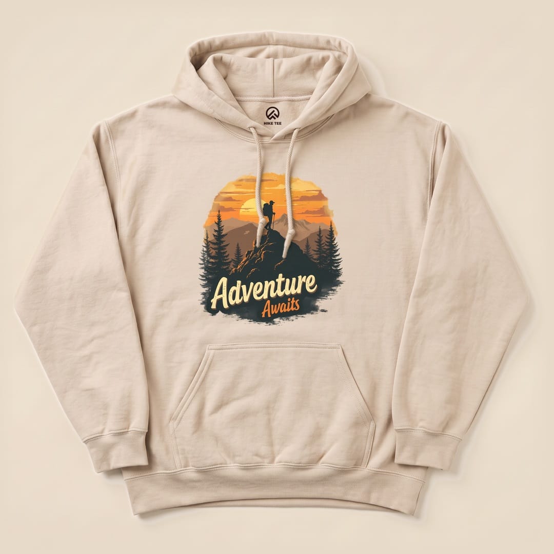 The Adventure Awaits Hoodie features a graphic of a hiker against a sunset backdrop with pine trees, printed on a beige hoodie.