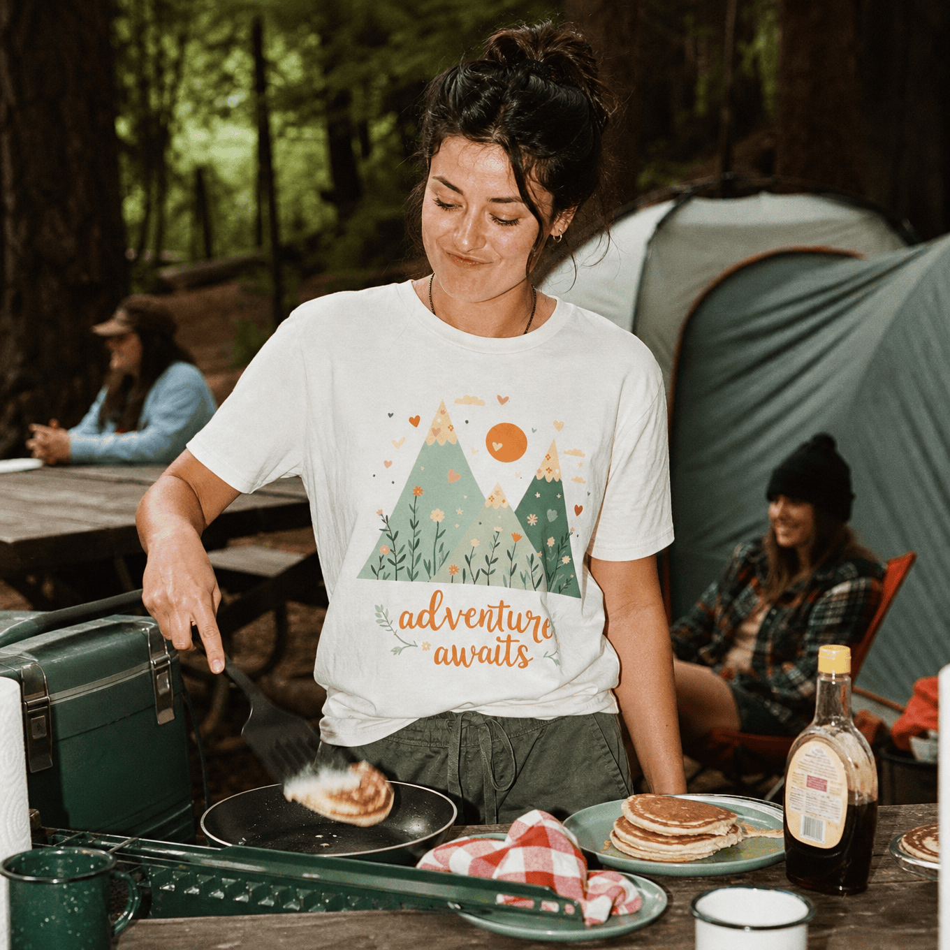A woman cooking pancakes outdoors is wearing a graphic t-shirt featuring a mountain design and the slogan "adventure awaits.