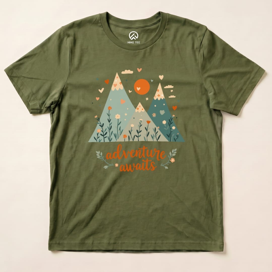 The Adventure Awaits Shirt features a graphic of colorful mountains with a heart and flowers, printed on an olive green t-shirt.
