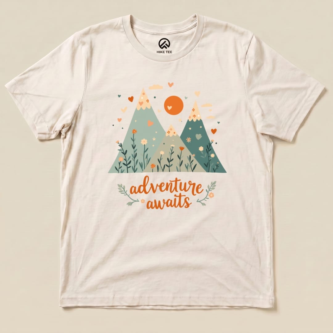 A graphic t-shirt featuring two mountains, flowers, and the phrase "adventure awaits" in a playful font.