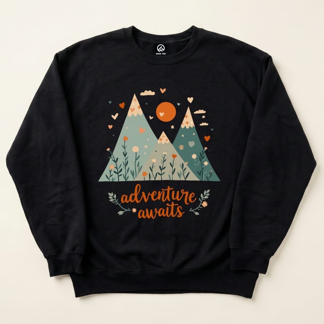 The Adventure Awaits Sweatshirt features a colorful graphic of mountains, a sun, and floral elements with the phrase "adventure awaits.
