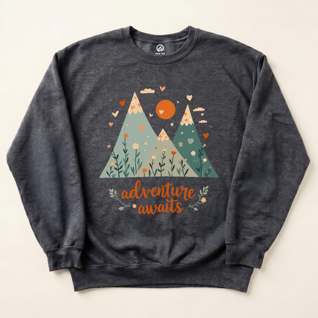 A dark gray "Adventure Awaits Sweatshirt" featuring a graphic of mountains, flowers, and a sun, with the text "adventure awaits" below.