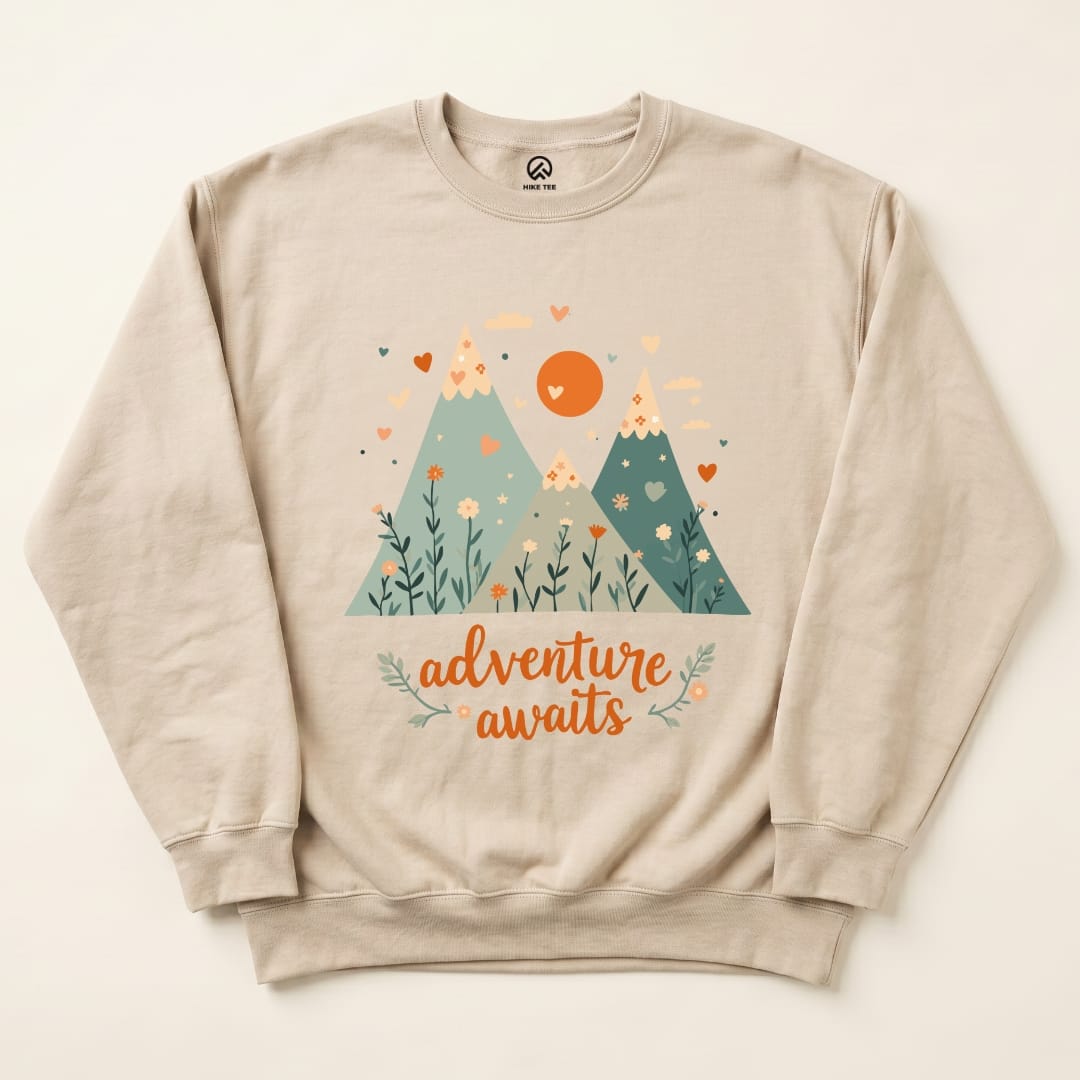 A beige "Adventure Awaits Sweatshirt" featuring a colorful mountain graphic with hearts, flowers, and the slogan below.