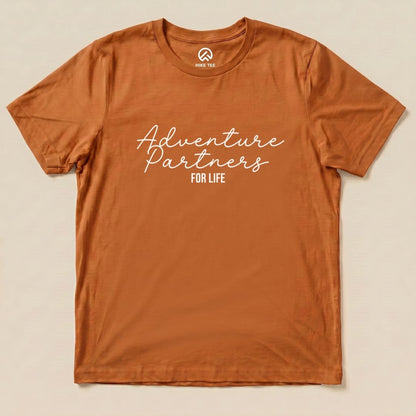A flat graphic t-shirt in rust color featuring the phrase "Adventure Partners for Life" in a stylish font.