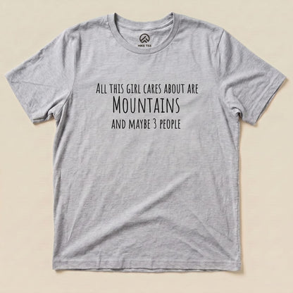 A flat graphic t-shirt labeled "All This Girl Cares Shirt," featuring the text: "All this girl cares about are Mountains and maybe 3 people.