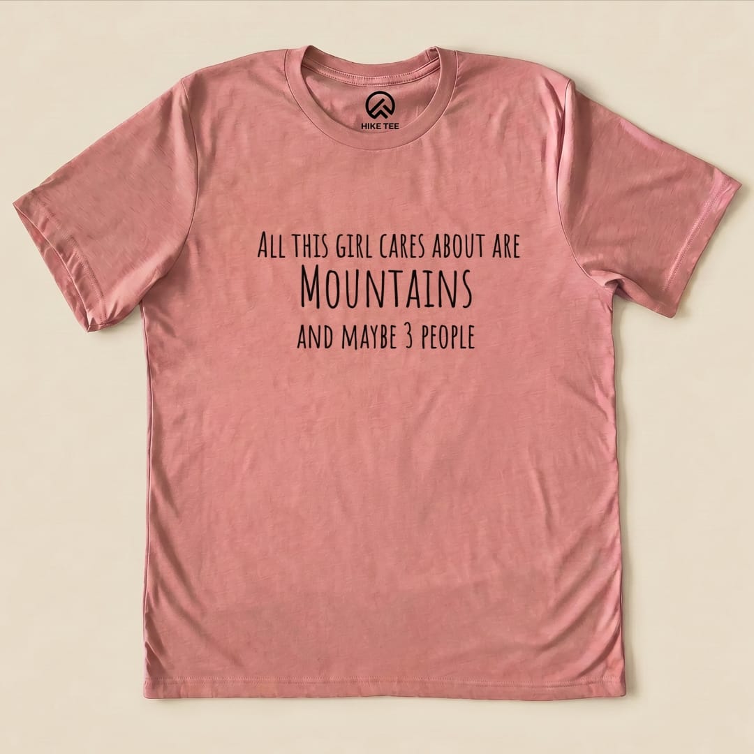 A pink graphic t-shirt featuring the phrase "All This Girl Cares About Are Mountains and Maybe 3 People.