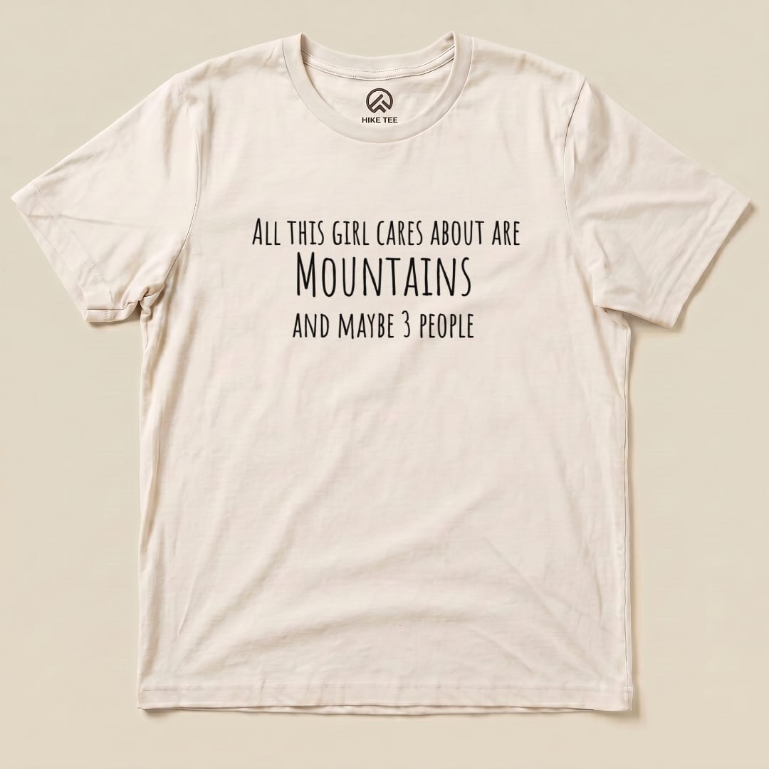 The image features a flat lay of the "All This Girl Cares Shirt," displaying the text "All this girl cares about are Mountains and maybe 3 people" in bold black font on a cream-colored graphic