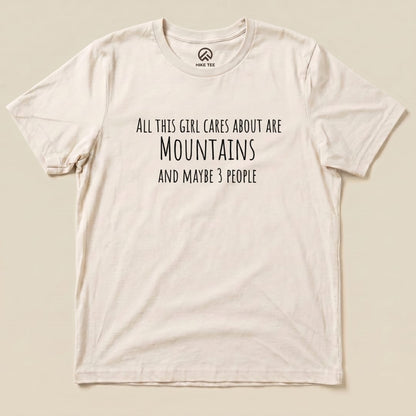 The image features a flat lay of the "All This Girl Cares Shirt," displaying the text "All this girl cares about are Mountains and maybe 3 people" in bold black font on a cream-colored graphic