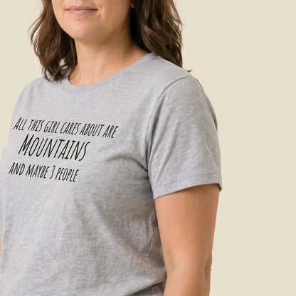 A woman wearing a graphic t-shirt that reads "All This Girl Cares About Are Mountains and Maybe 3 People" in black text.