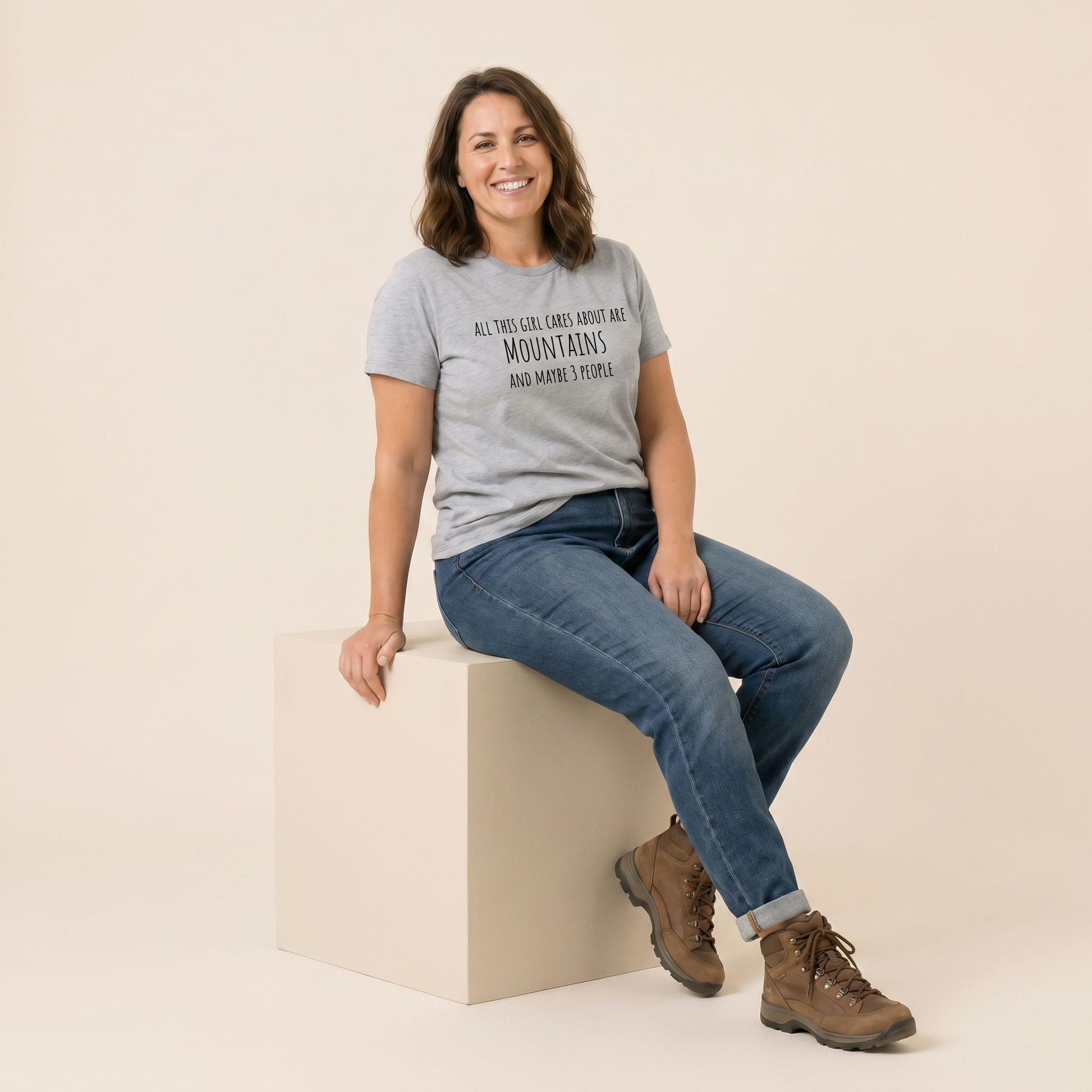A woman sits on a cube wearing a gray graphic t-shirt that reads "All This Girl Cares About Are Mountains and Maybe 3 People.