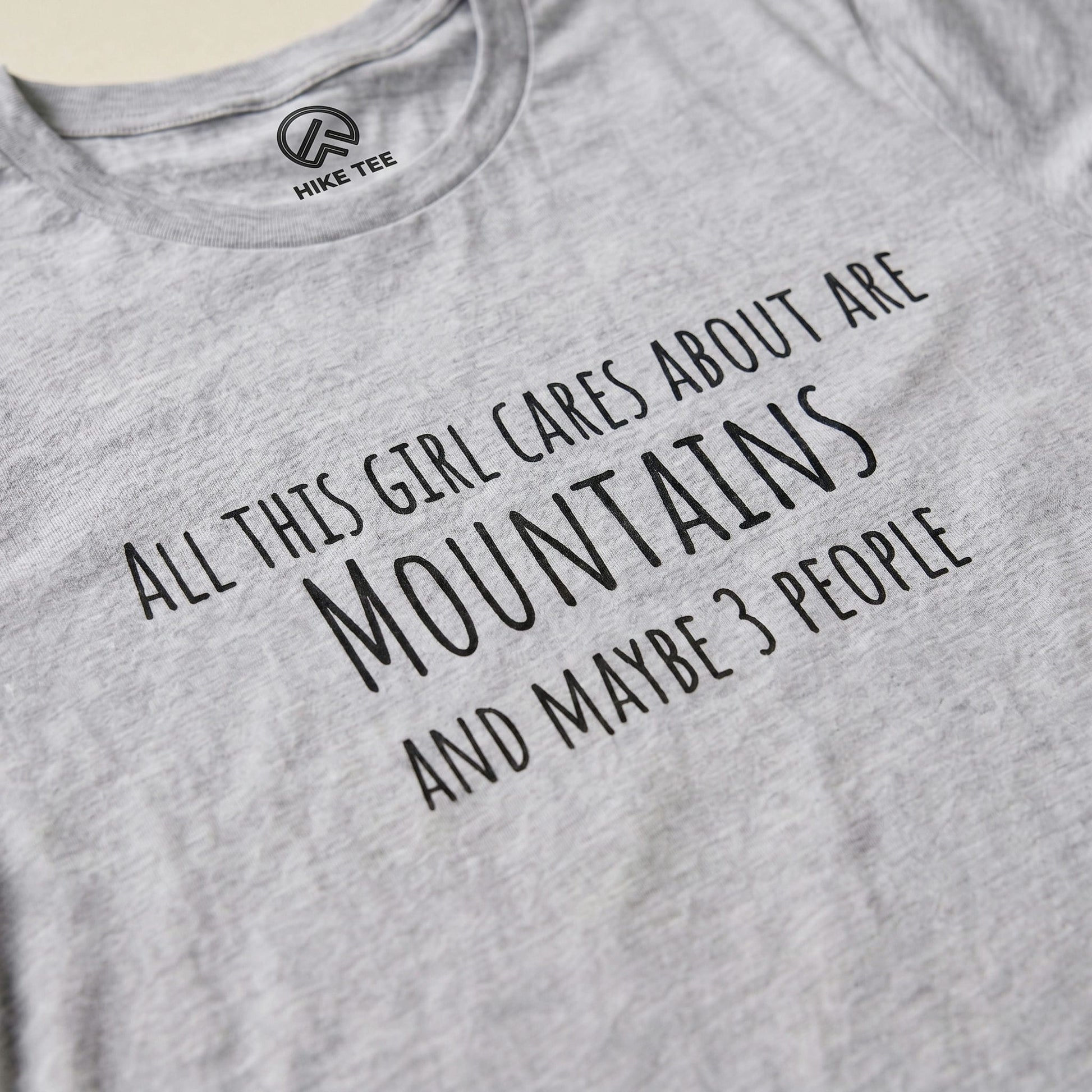 The All This Girl Cares Shirt features bold text stating, "All this girl cares about are MOUNTAINS and maybe 3 PEOPLE" on a gray graphic t-shirt.