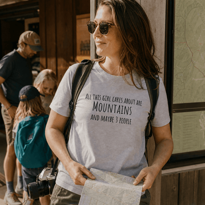 A woman stands outdoors, wearing a graphic t-shirt that reads "All This Girl Cares About Are Mountains and Maybe 3 People.