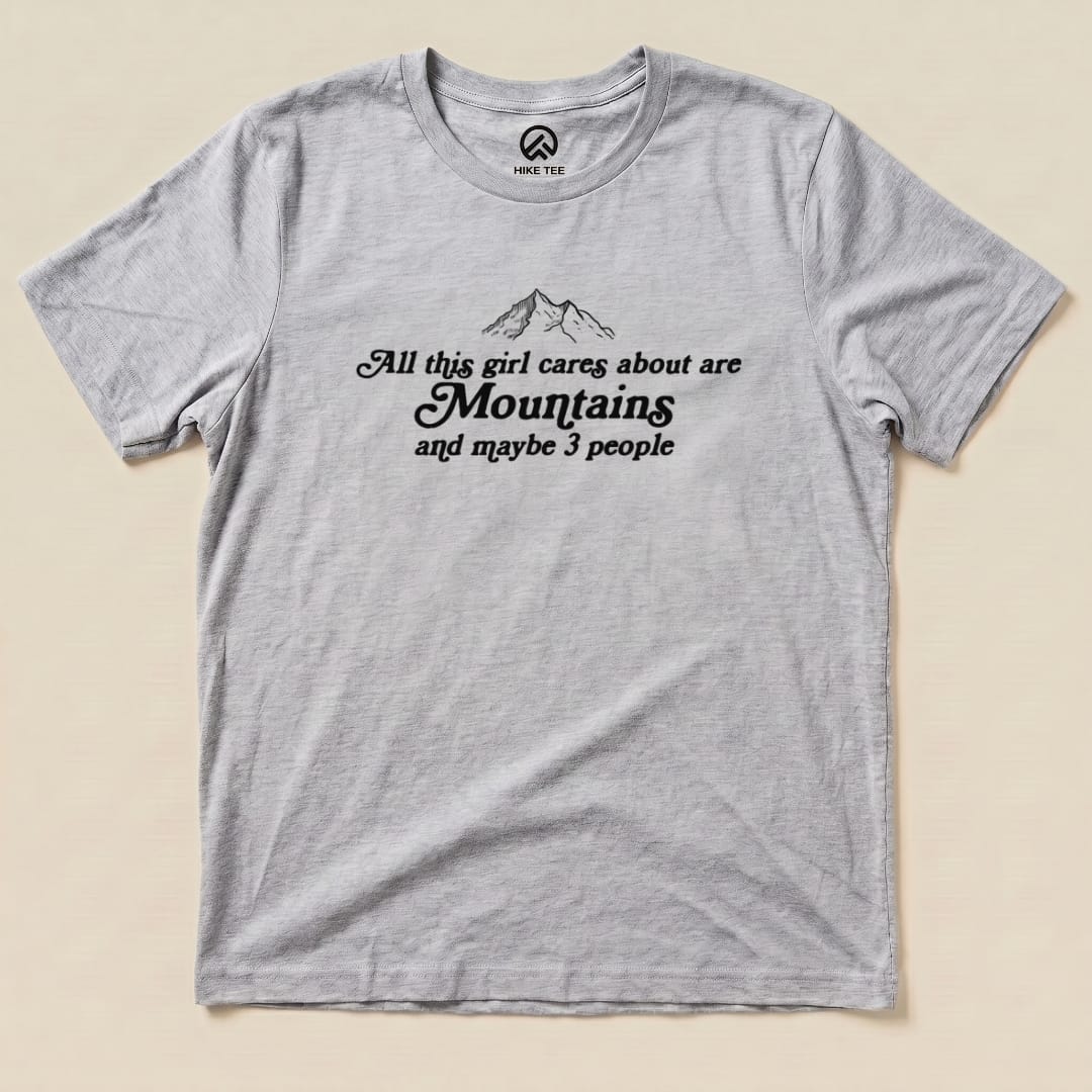 Gray graphic t-shirt featuring the text "All this girl cares about are Mountains and maybe 3 people" with mountain illustrations.