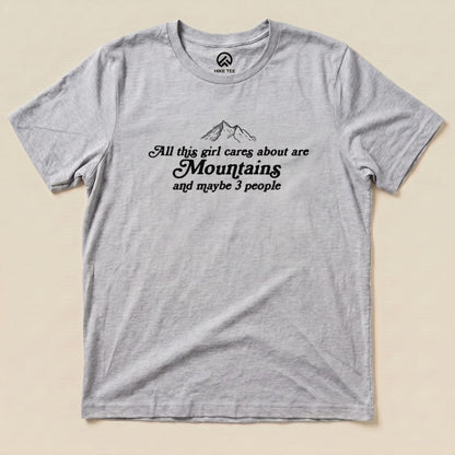Gray graphic t-shirt featuring the text "All this girl cares about are Mountains and maybe 3 people" with mountain illustrations.