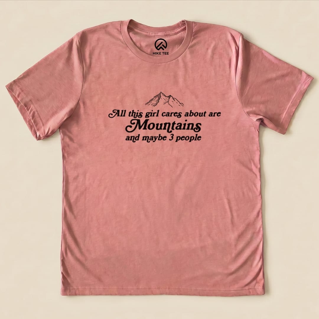 A pink graphic t-shirt featuring the phrase "All This Girl Cares About Are Mountains and Maybe 3 People" with mountain illustrations.